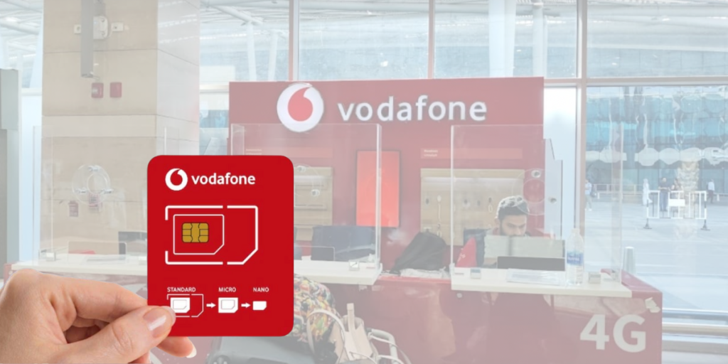 A hand holding a Vodafone physical SIM card in front of a Vodafone kiosk at Cairo International Airport